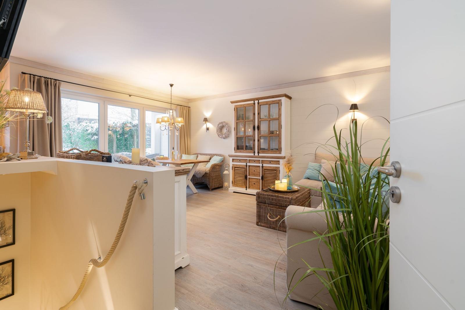 Wohnbereich mit Sofa, Tisch und Holzschränken. Große Fenster und Pflanzen sorgen für viel Licht.