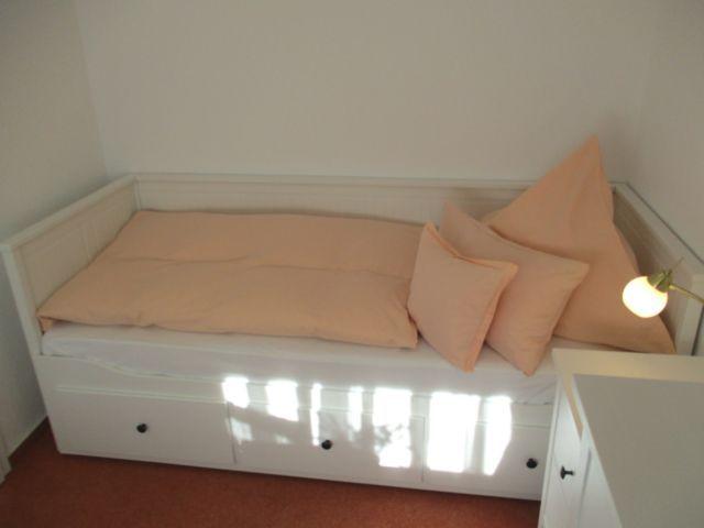 Ein Bett mit orangefarbenen Kissen und Matratze. Unterseite mit Schubladen.