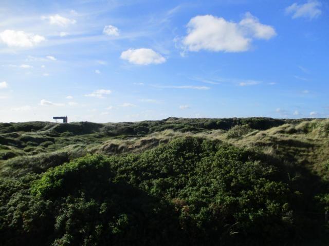 Weitläufige Dünenlandschaft unter blauem Himmel mit vereinzelten Wolken und einem Holzgestell im Hintergrund.
