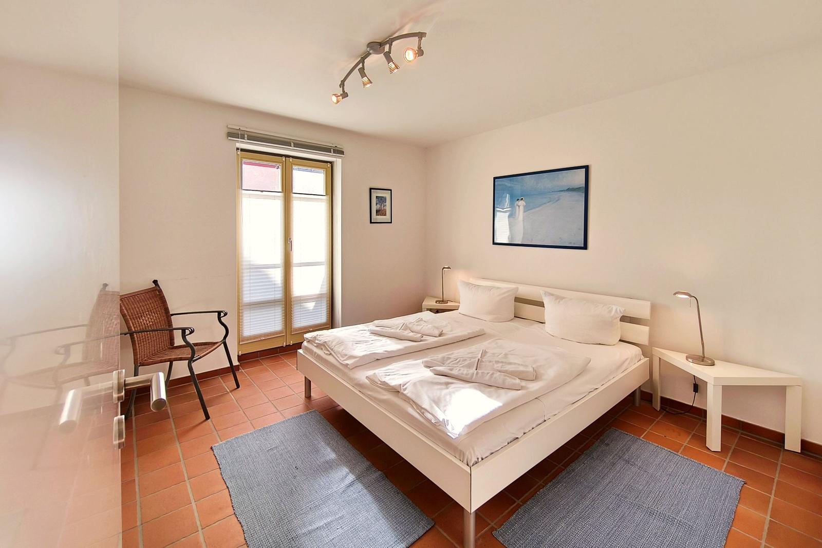 Schlafzimmer mit Doppelbett, zwei Nachttischen und Stuhl vor Fenster.