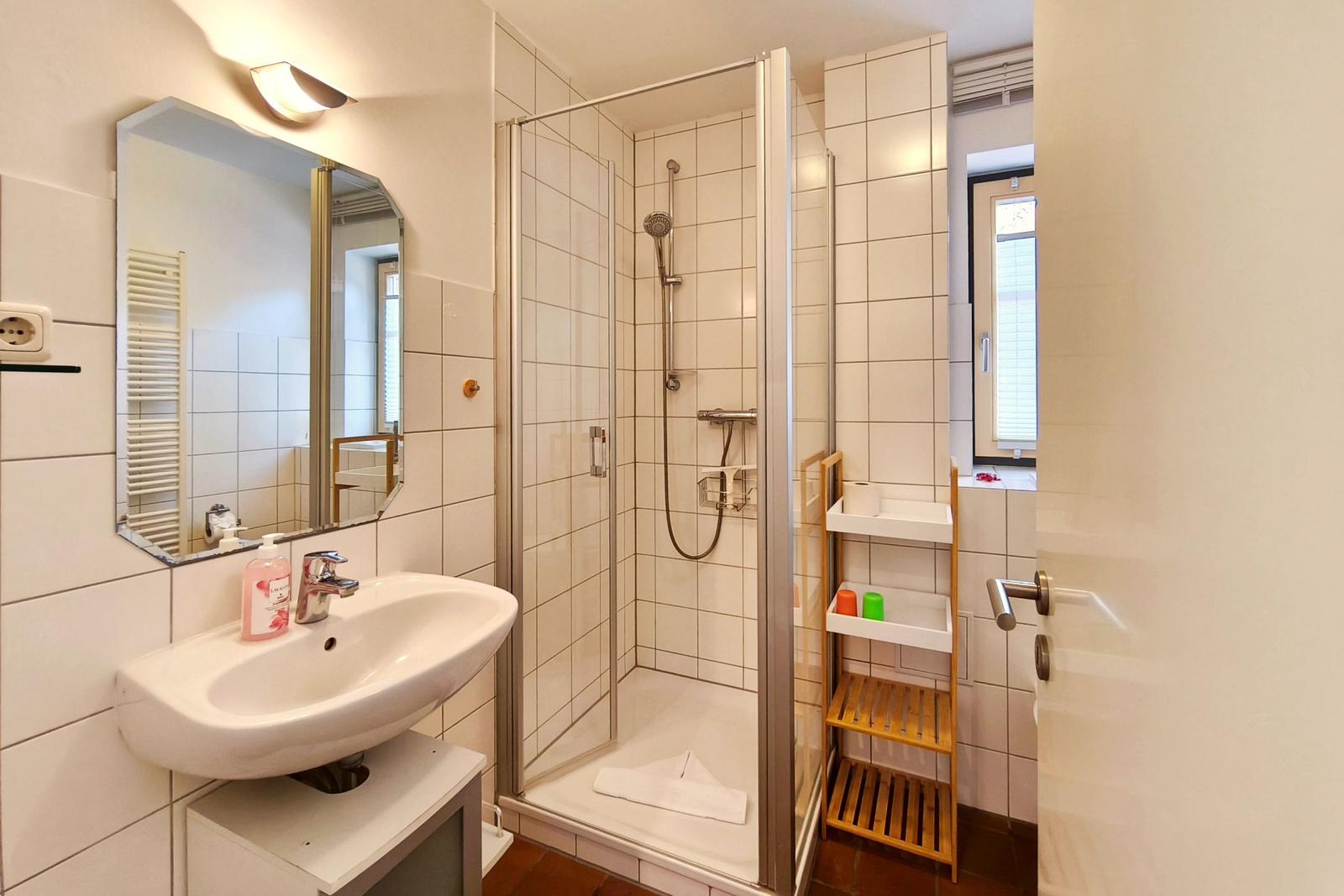 Badezimmer mit Dusche, Waschbecken und Spiegel. Holzregal und Fenster sichtbar.