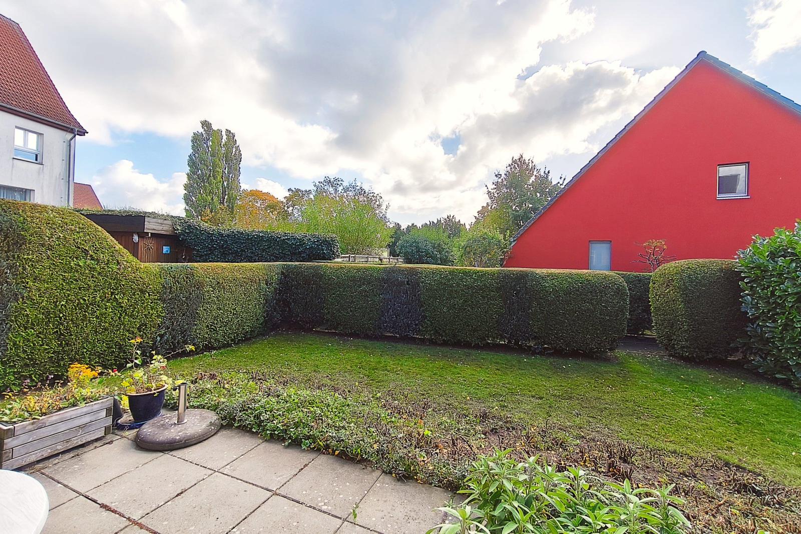 Roter Gartenbereich mit Rasen, Hecken und Pflanzen. Hintergrund mit Bäumen und Häusern.