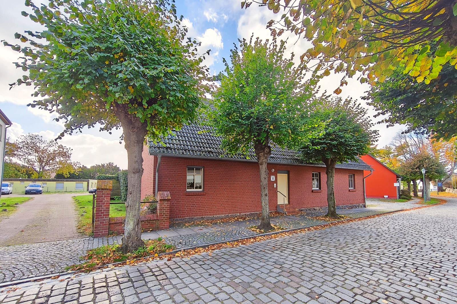 Rotes Backsteingebäude mit Eingangstür und Fenstern unter Bäumen auf Kiesweg.