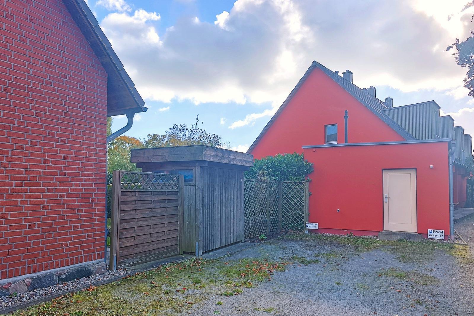 Rotes Gebäude mit Eingangstür und Holztor. Gras und Kies vor dem Haus.