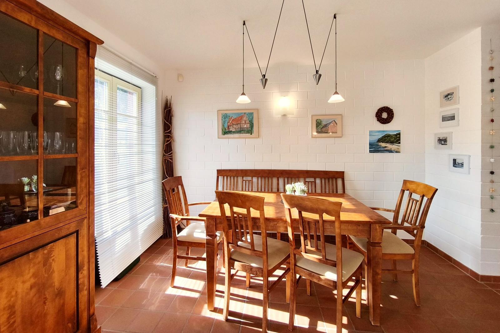 Esszimmer mit Holztisch, Stühlen und Wandbildern. Fenster mit Jalousien und Hängelampen.