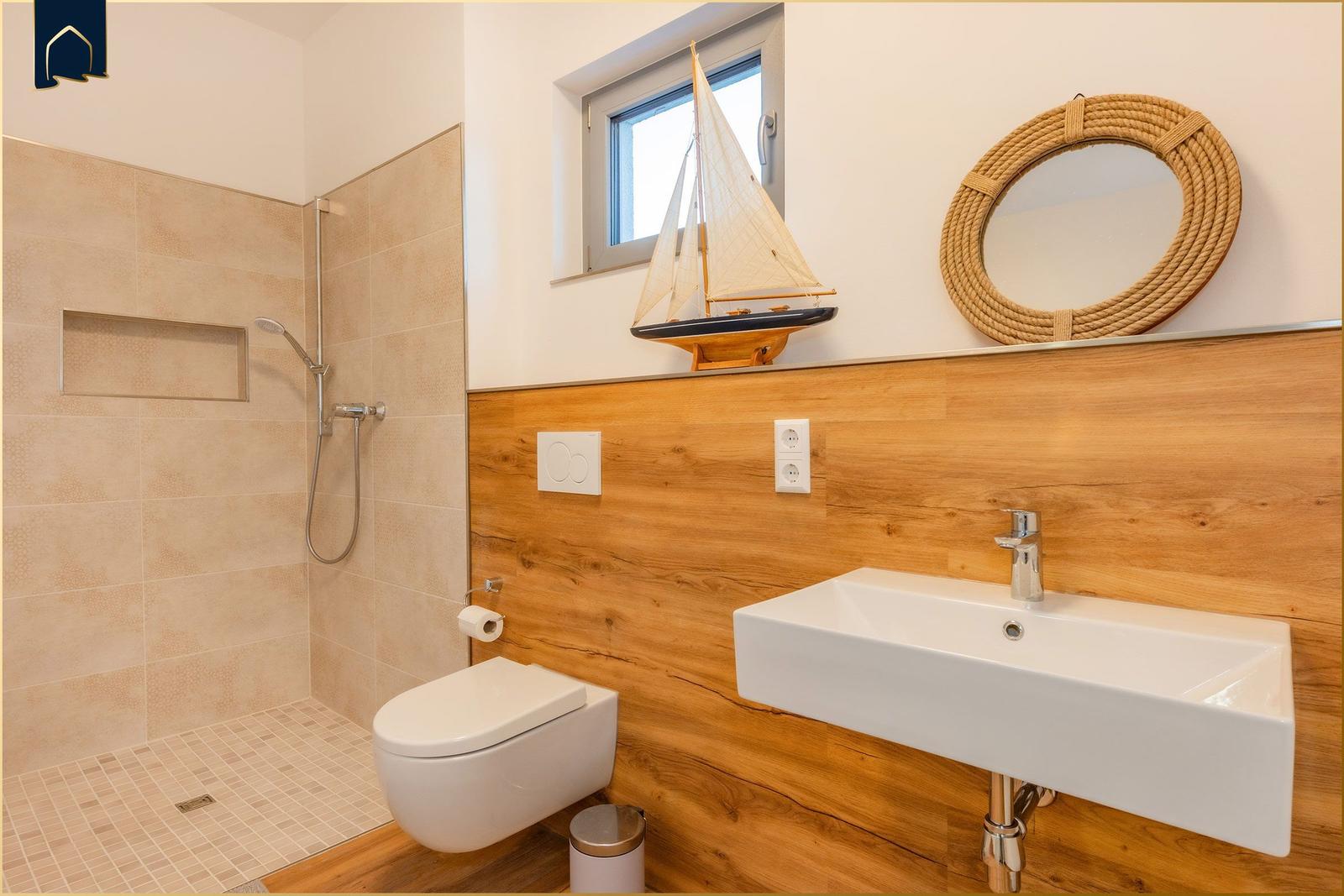 Badezimmer mit Dusche, Toilette und Waschbecken. Holzwand, Spiegel und Segelboot-Dekoration.