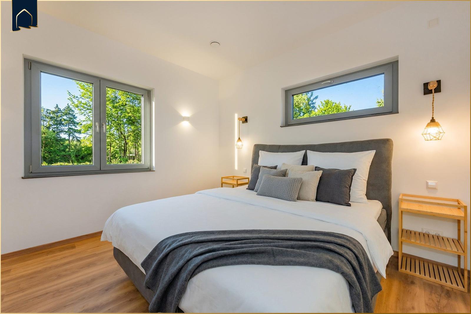 Schlafzimmer mit Bett, Fenstern und Holzboden. Blick auf grüne Bäume.