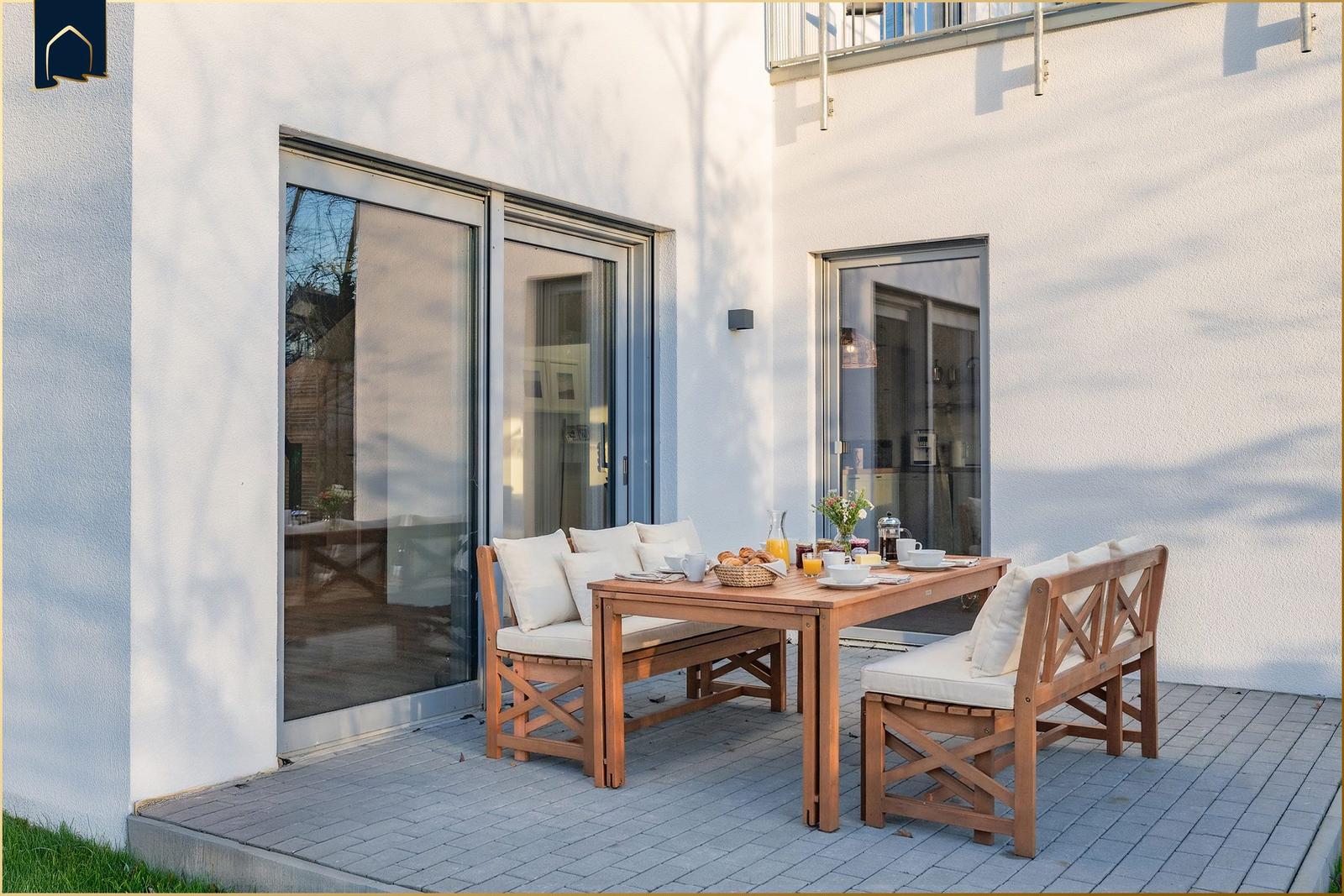 Terrasse mit Holztisch und Bänken, darauf Frühstücksgedecke. Hintergrund: weiße Wand und Glastüren.