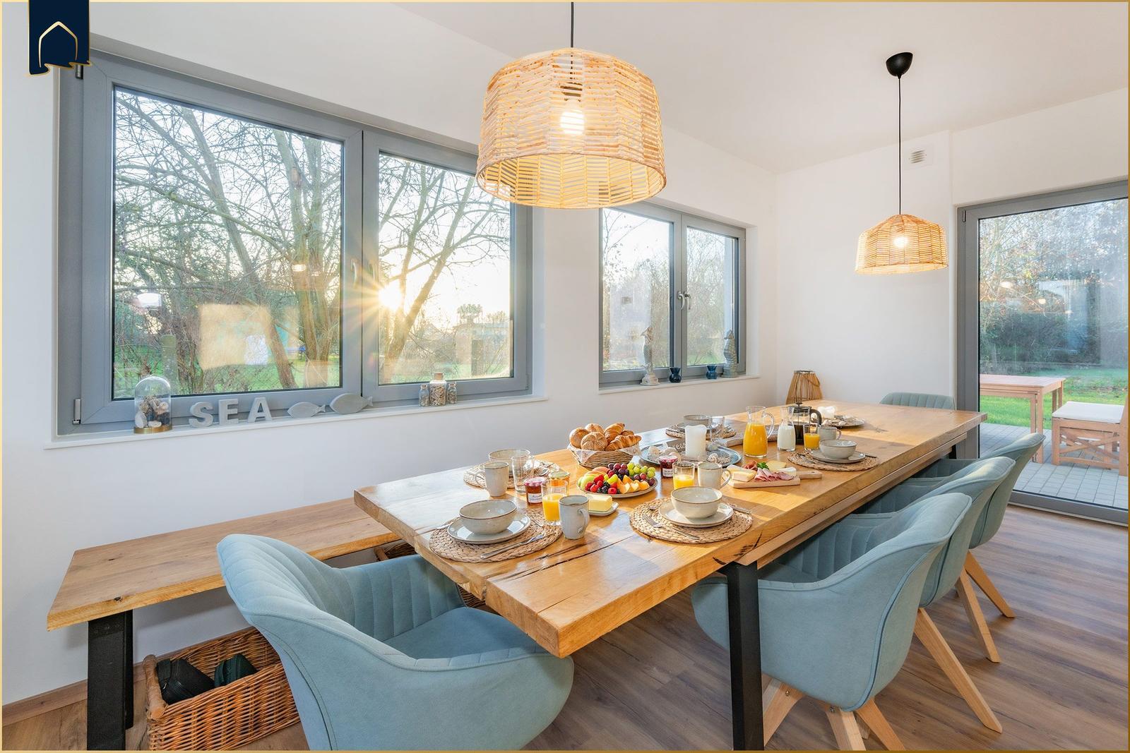 Esszimmer mit langem Holztisch, blauen Stühlen und großen Fenstern mit Blick auf Garten.