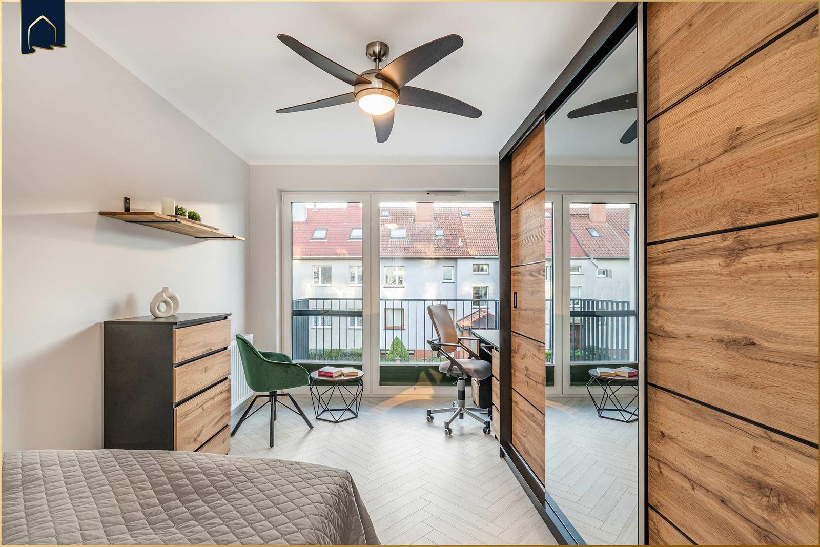 Schlafzimmer mit Balkon, Schreibtisch und Holzschrank. Deckenventilator und Hocker sichtbar.