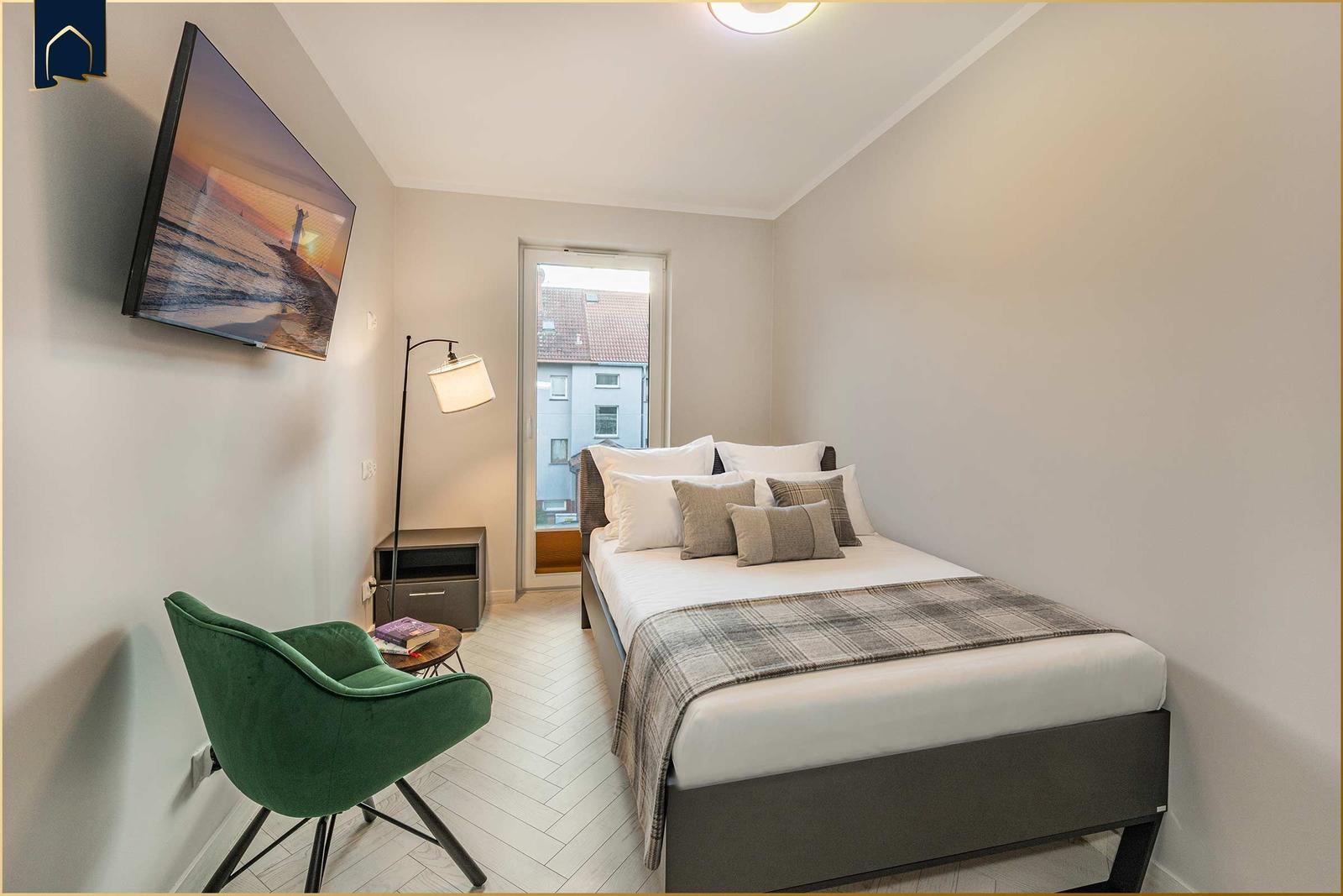 Schlafzimmer mit Bett, grüner Sessel und Fernseher an der Wand.