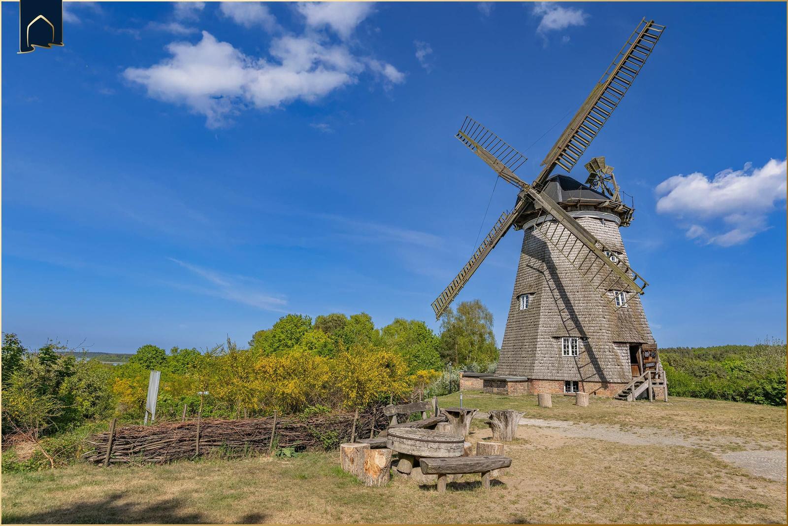 Holzmühle mit Sitzgruppe auf grünem Grund unter blauem Himmel.