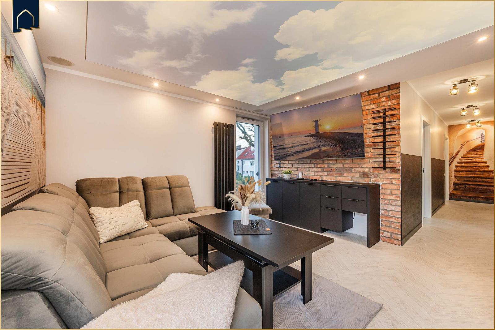 Wohnzimmer mit Sofa, Couchtisch und Fernseher an Backsteinwand. Decke mit Wolkenmotiv.