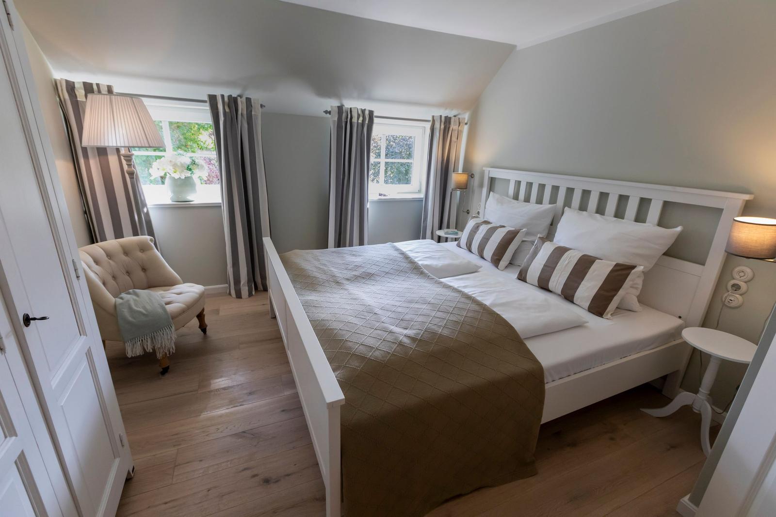 Ein Schlafzimmer mit weißem Bett, Holzboden und zwei Fenstern mit Vorhängen.