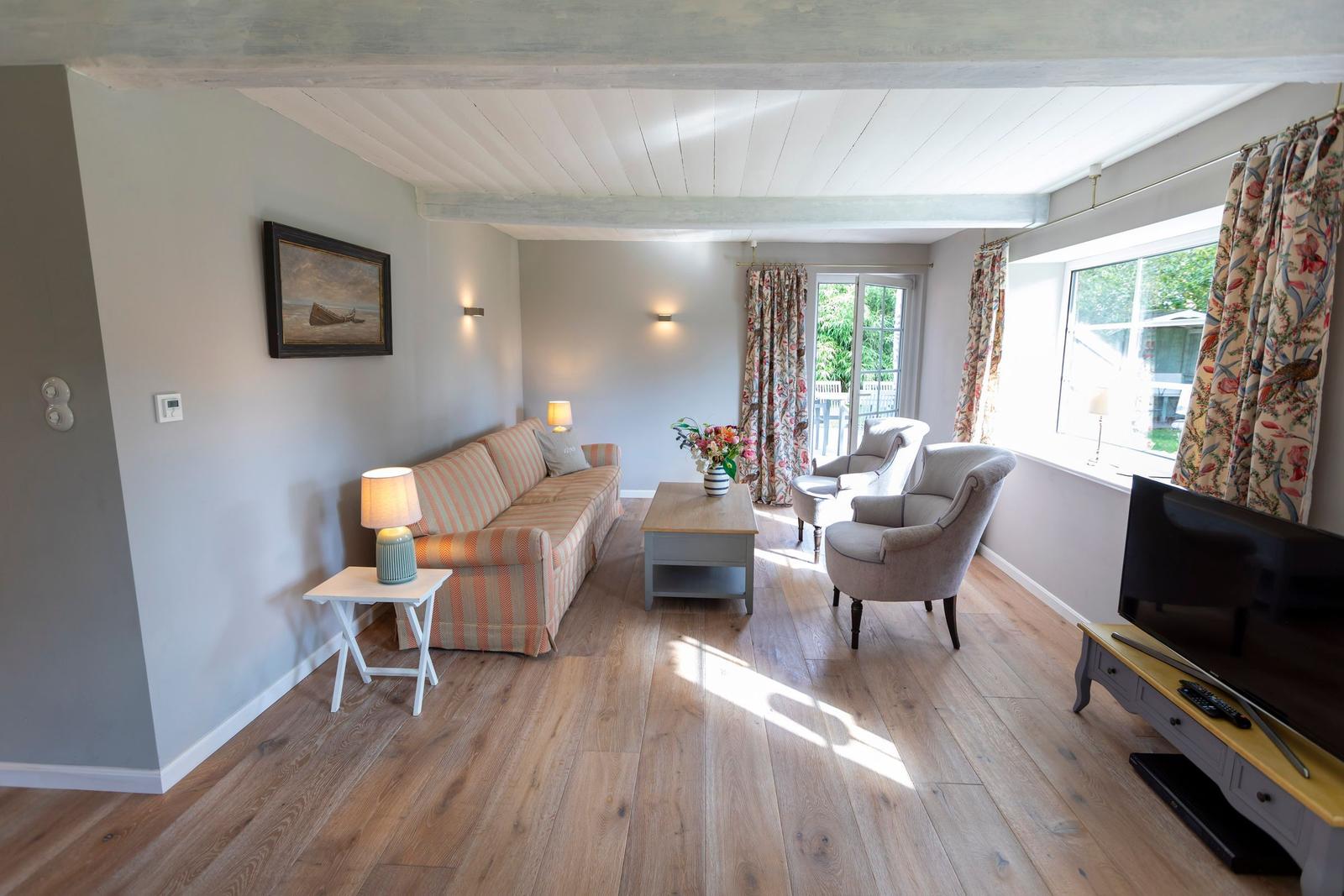 Wohnzimmer mit Sofa, Sesseln, Couchtisch und Fernseher. Holzboden und große Fenster.