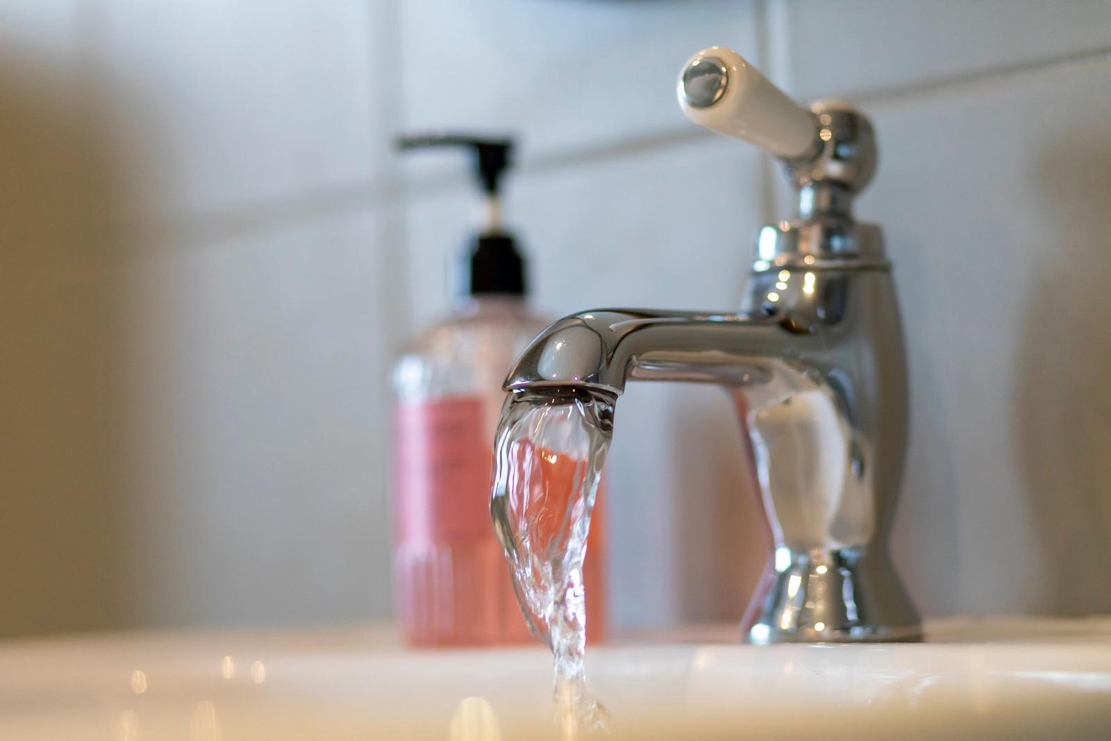 Ein Wasserhahn gießt Wasser in ein Waschbecken. Im Hintergrund steht eine Seifenspenderflasche.
