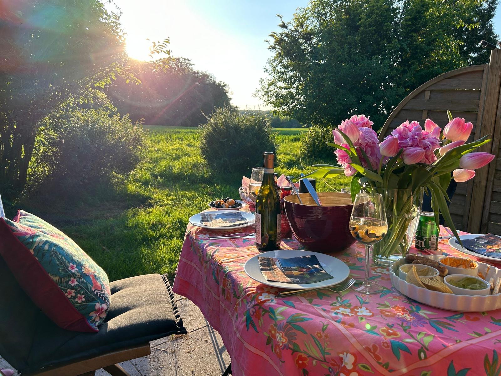 Tisch im Garten mit Blumen, Wein und Speisen bei Sonnenuntergang.