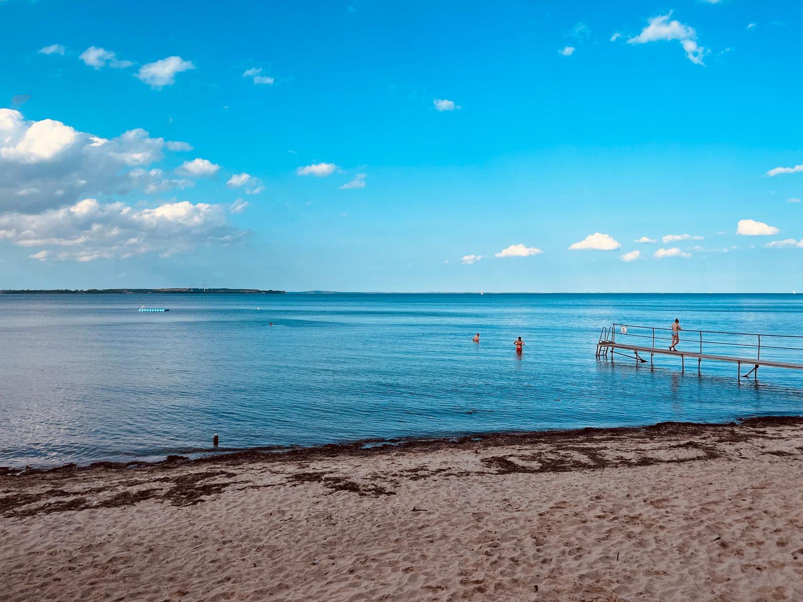 Strand mit Steg und Badegästen im klaren Meer unter blauem Himmel.