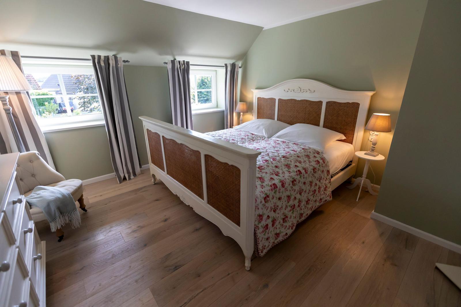 Schlafzimmer mit Bett, Nachttisch, Stuhl und Fenstern mit Vorhängen.