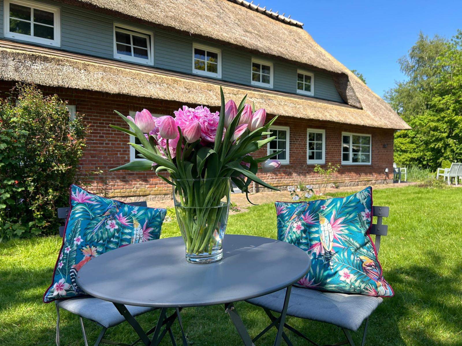 Runder Tisch mit Blumen und Kissen im Garten vor einem Strohdachhaus.
