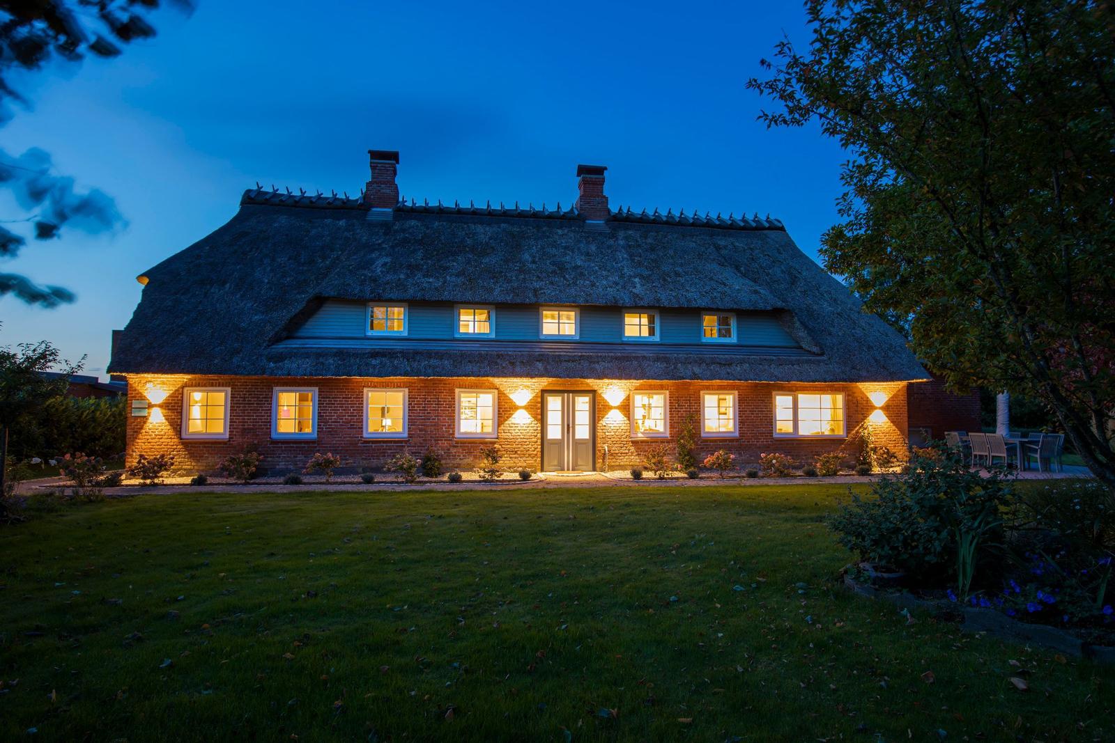 Haus mit strohgedecktem Dach und beleuchteten Fenstern bei Dämmerung.