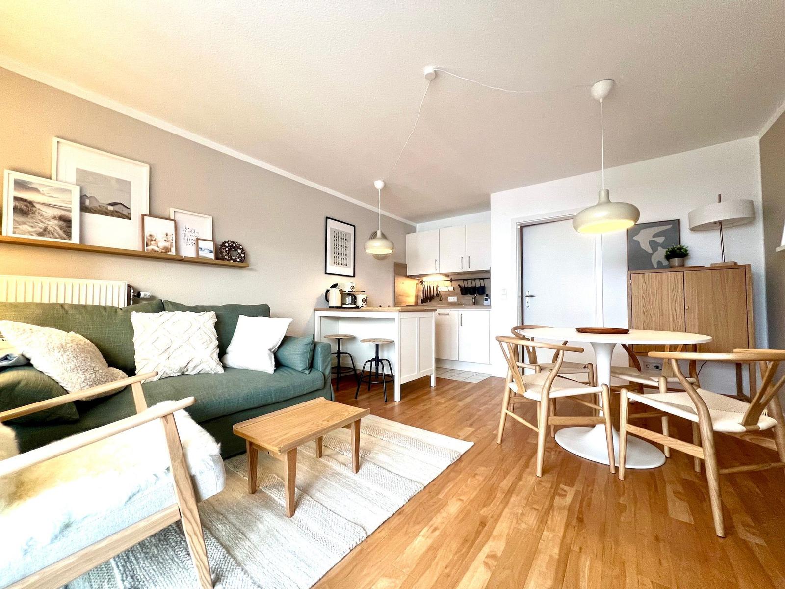 Wohnzimmer mit Sofa, Esstisch und Küche im offenen Raum. Holzfußboden und weiße Möbel.