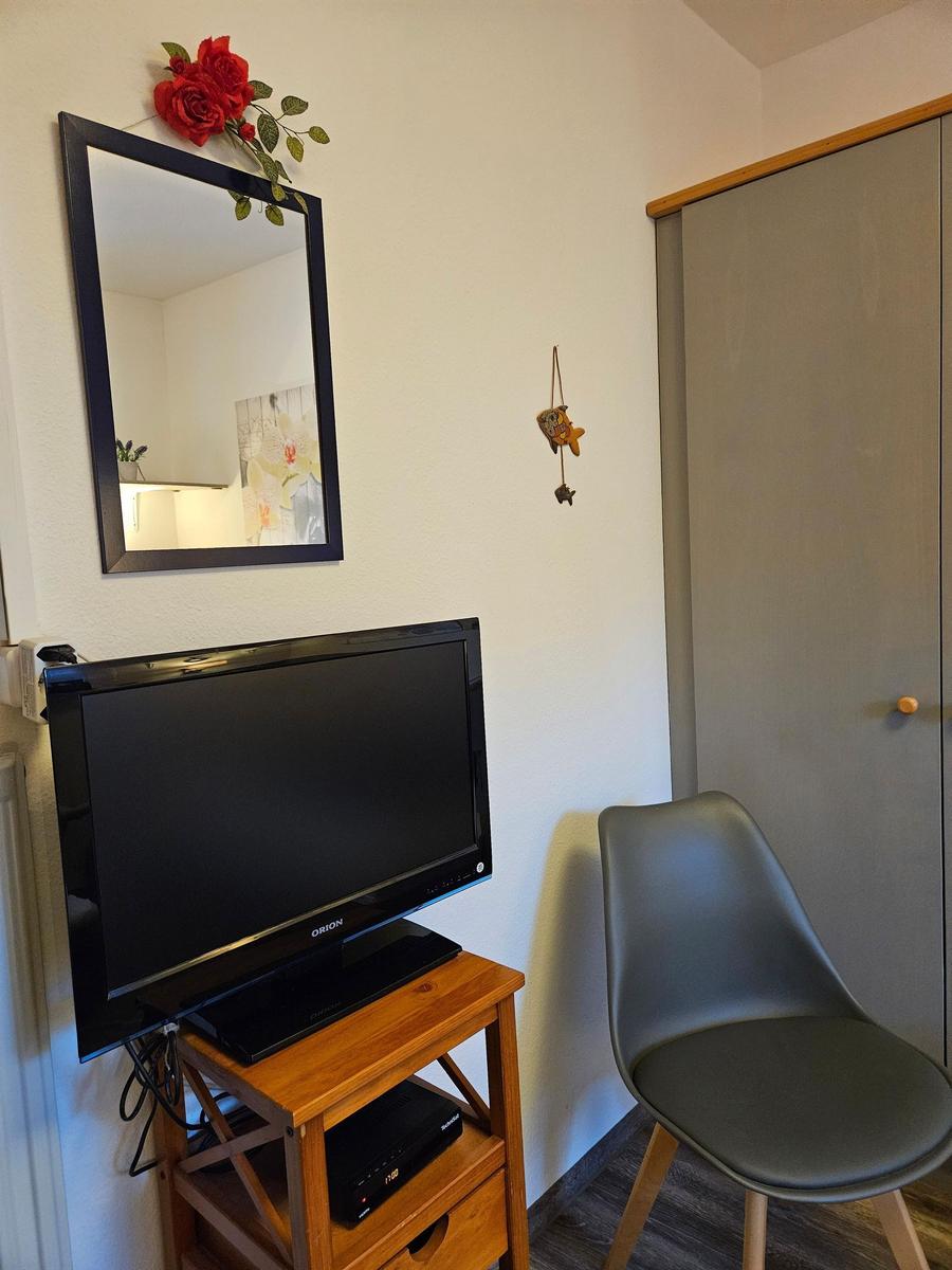 TV auf Holzstand, daneben Stuhl und Kleiderschrank. Spiegel mit roter Rose oben.