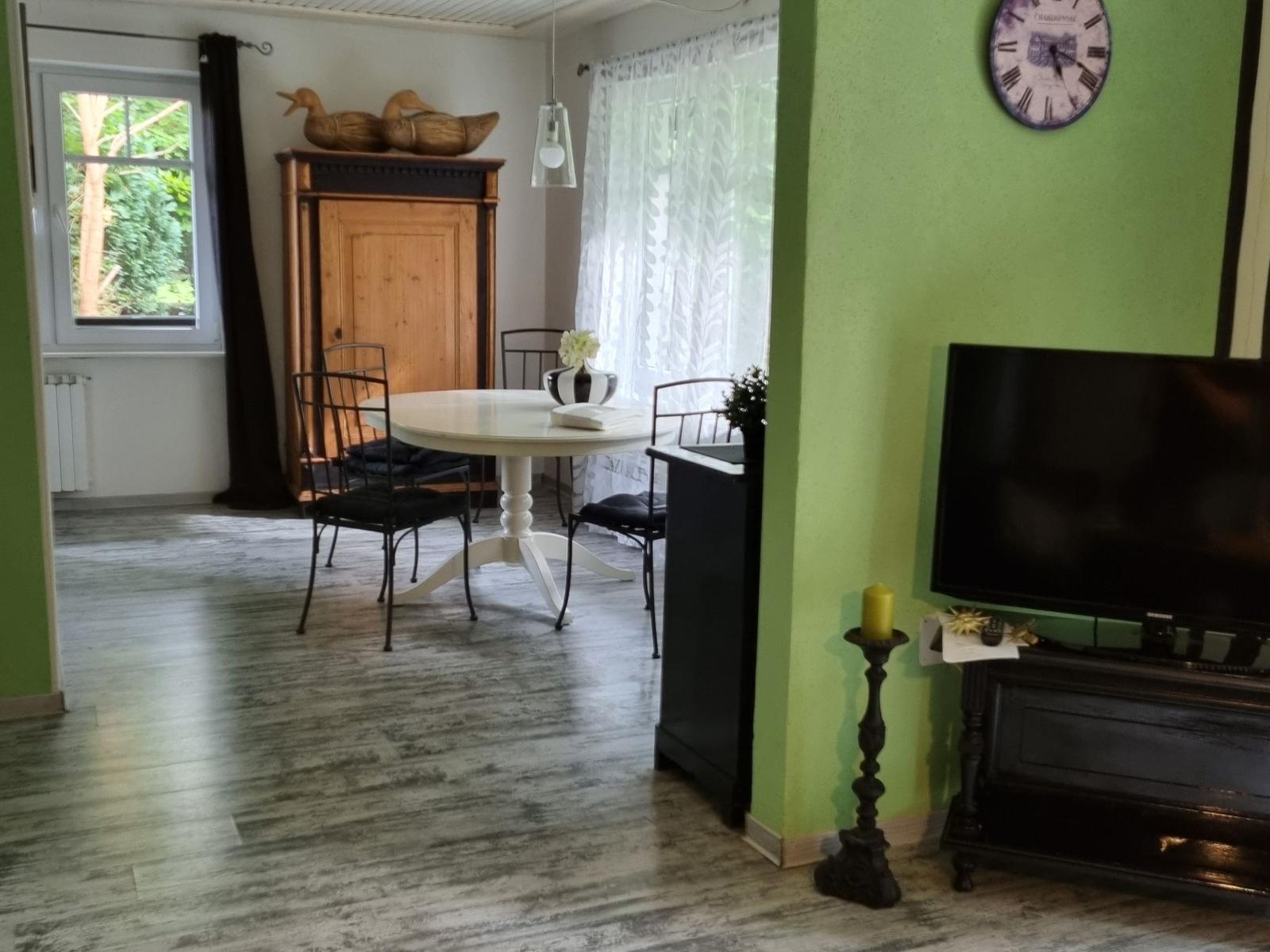 Dining area with round table and chairs. TV and wall clock are visible.