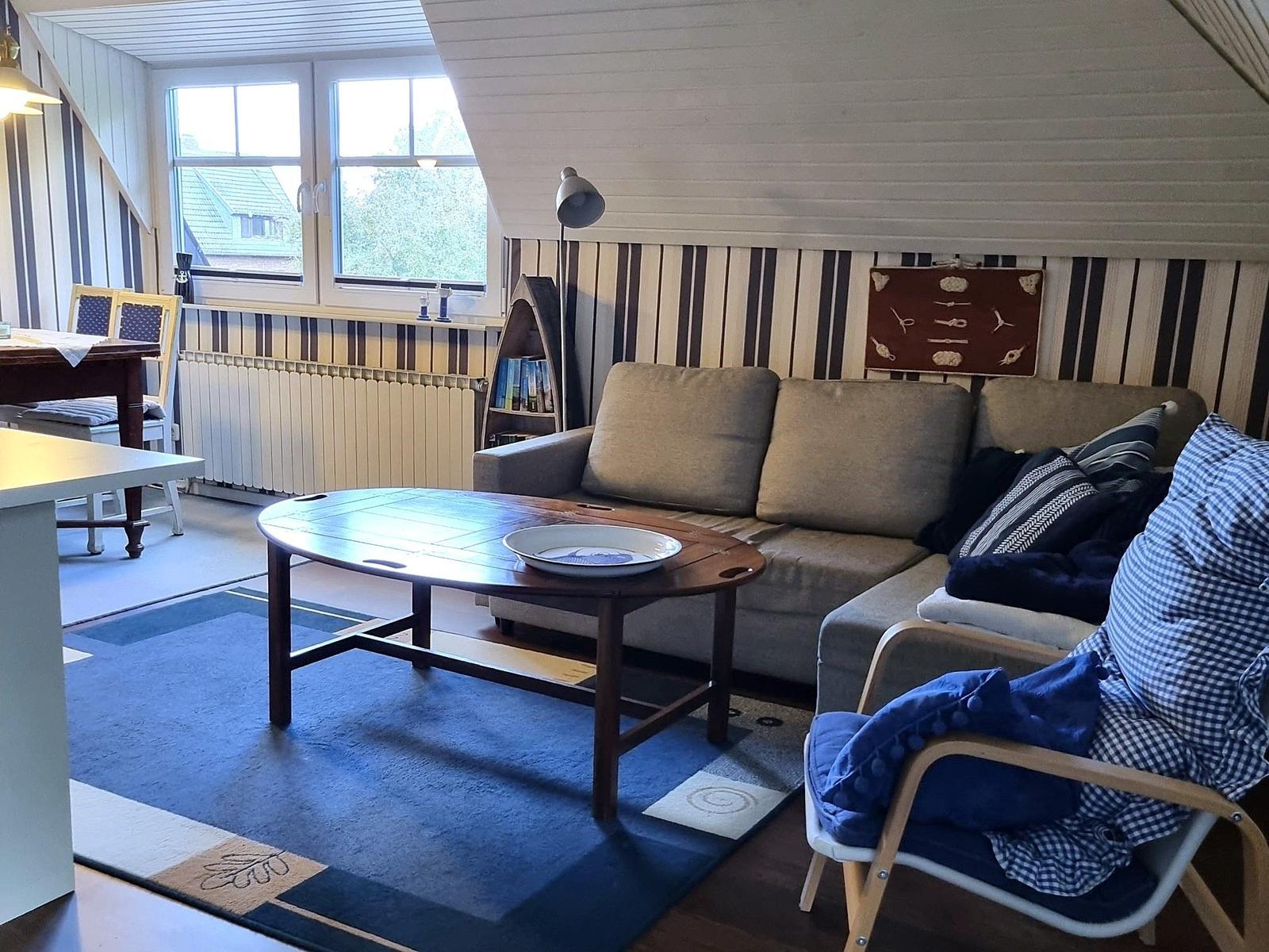 Living room with sofa, table, and chair. Window and wall paneling visible.