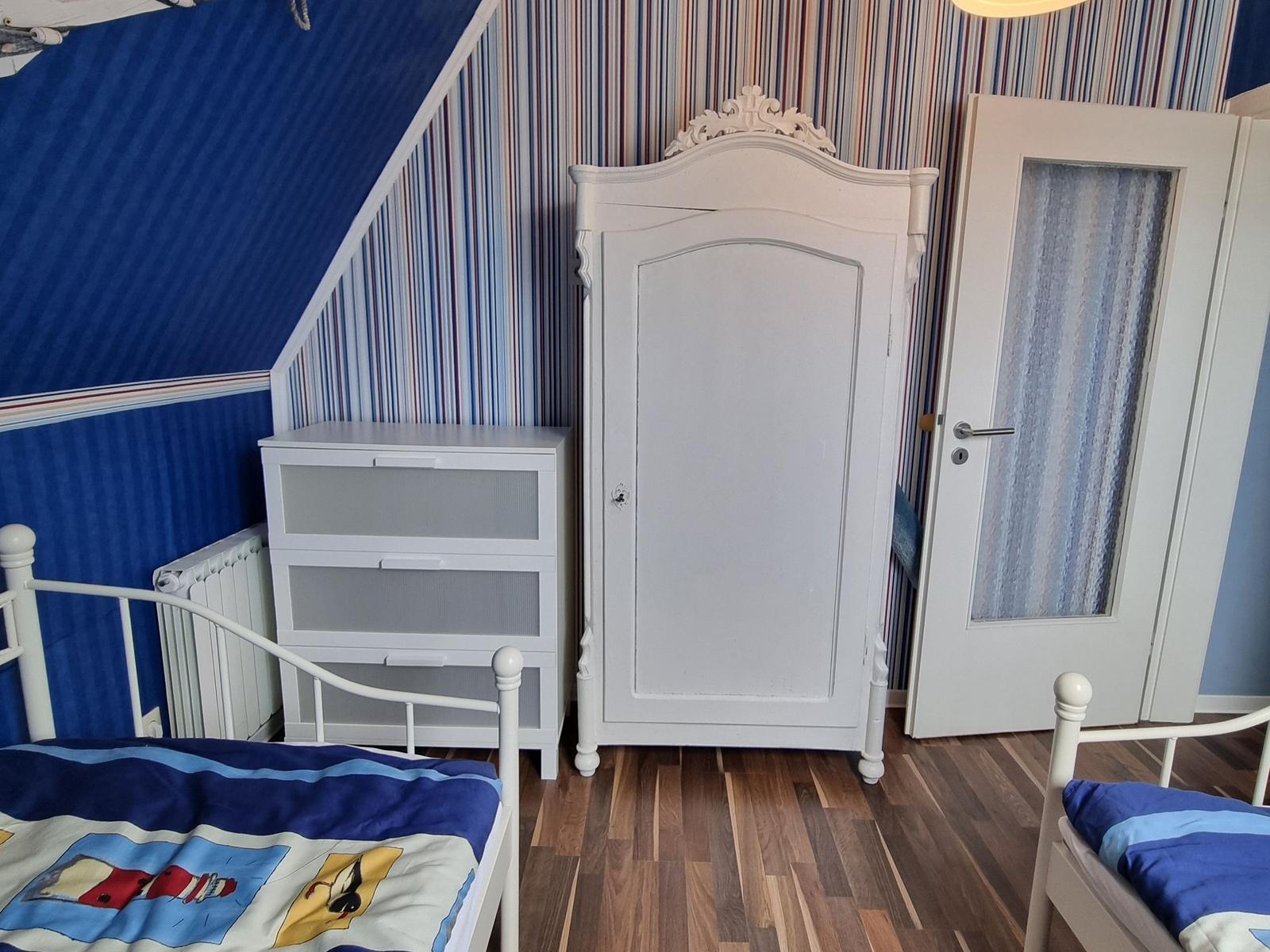 Room with two beds, wardrobe and chest. Striped wall and wooden floor.
