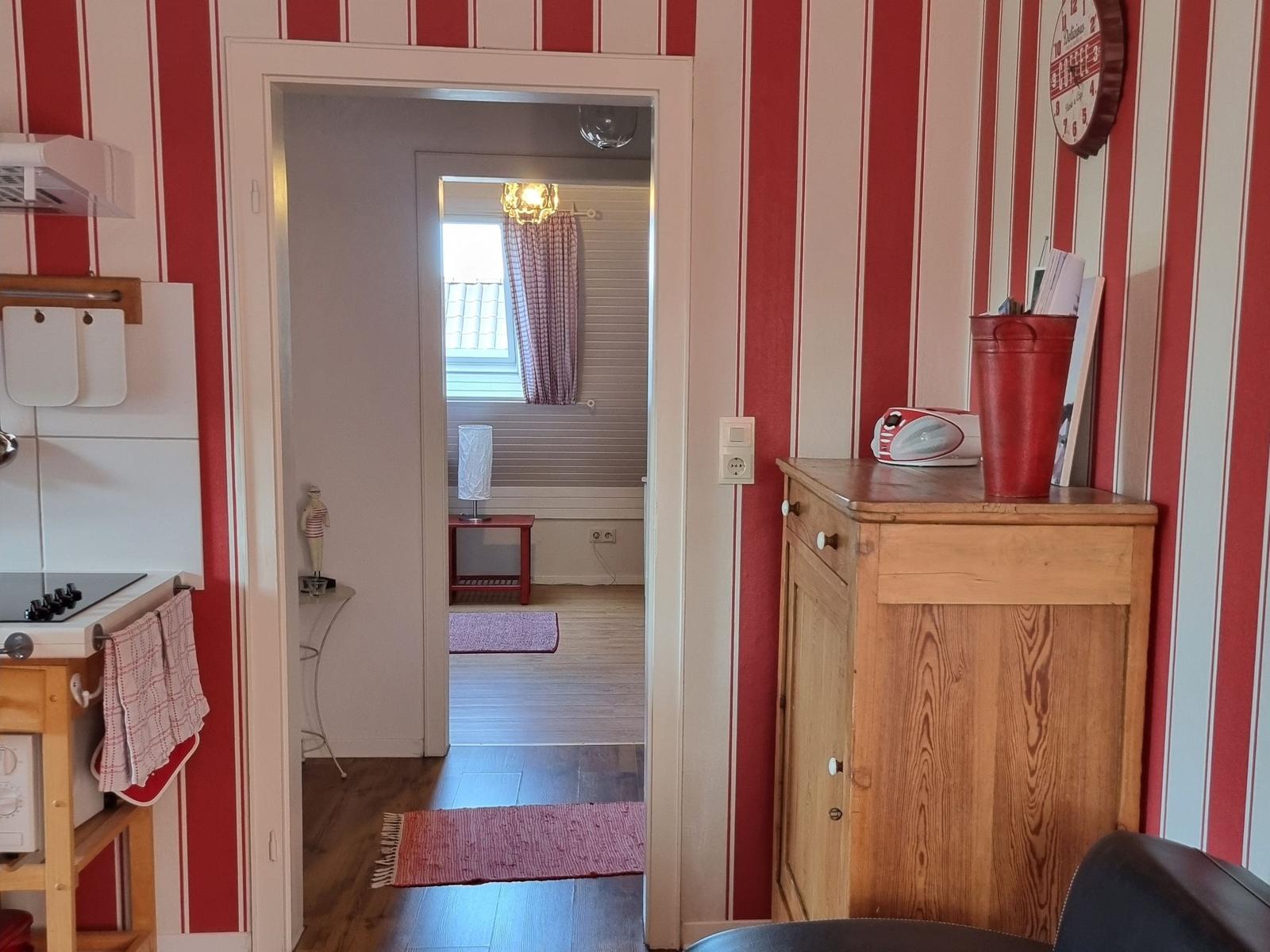 Kitchen with view into bedroom. Red striped wall, wooden furniture, and window with curtain.