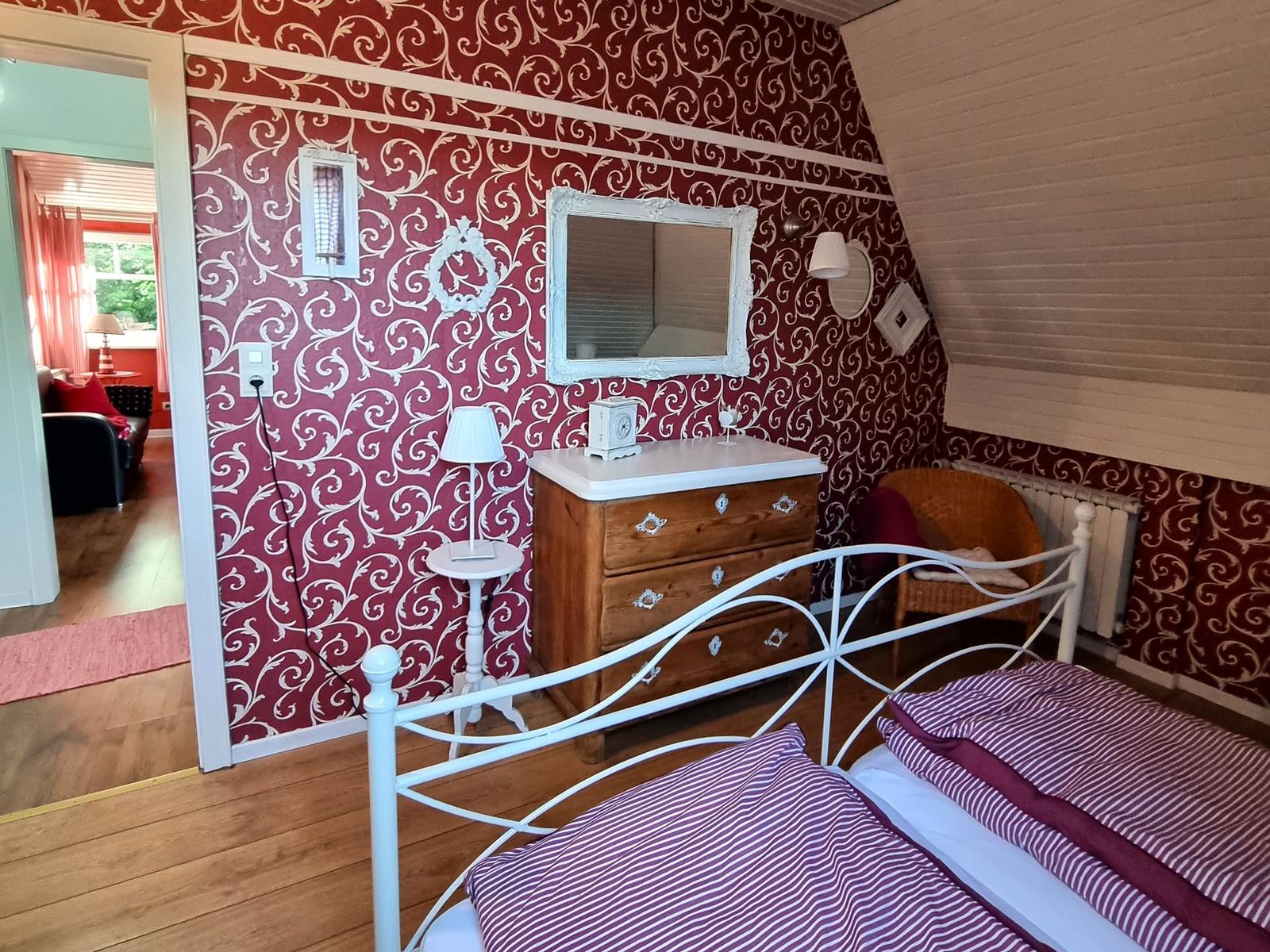 Bedroom with red patterned walls, white bed frame, and wooden dresser.