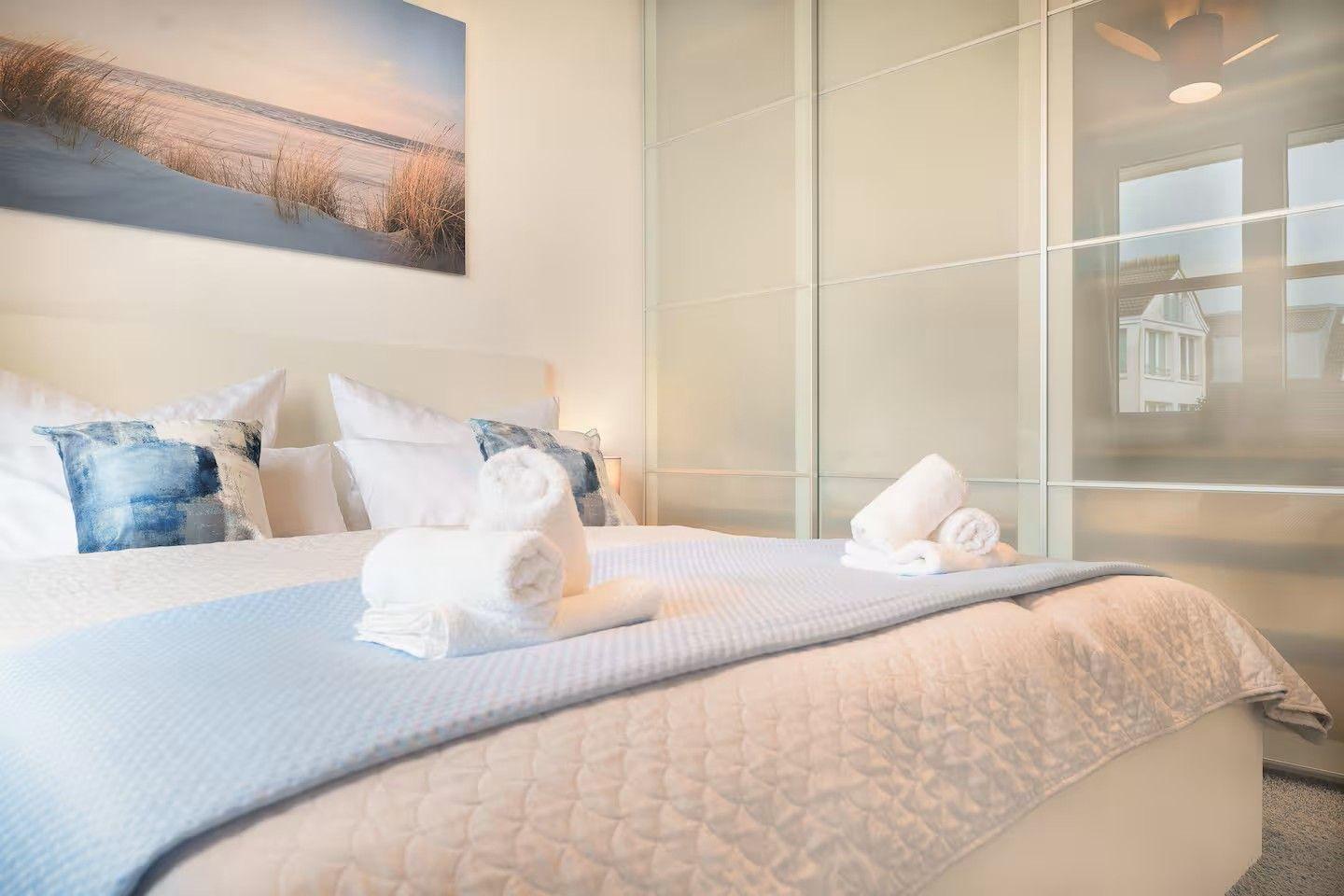 Schlafzimmer mit Bett, Kissen und Handtüchern. Wandbild und Kleiderschrank sichtbar.