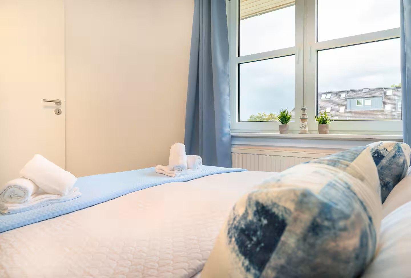 Schlafzimmer mit Bett, Fenster und Vorhängen. Auf dem Bett liegen Handtücher.