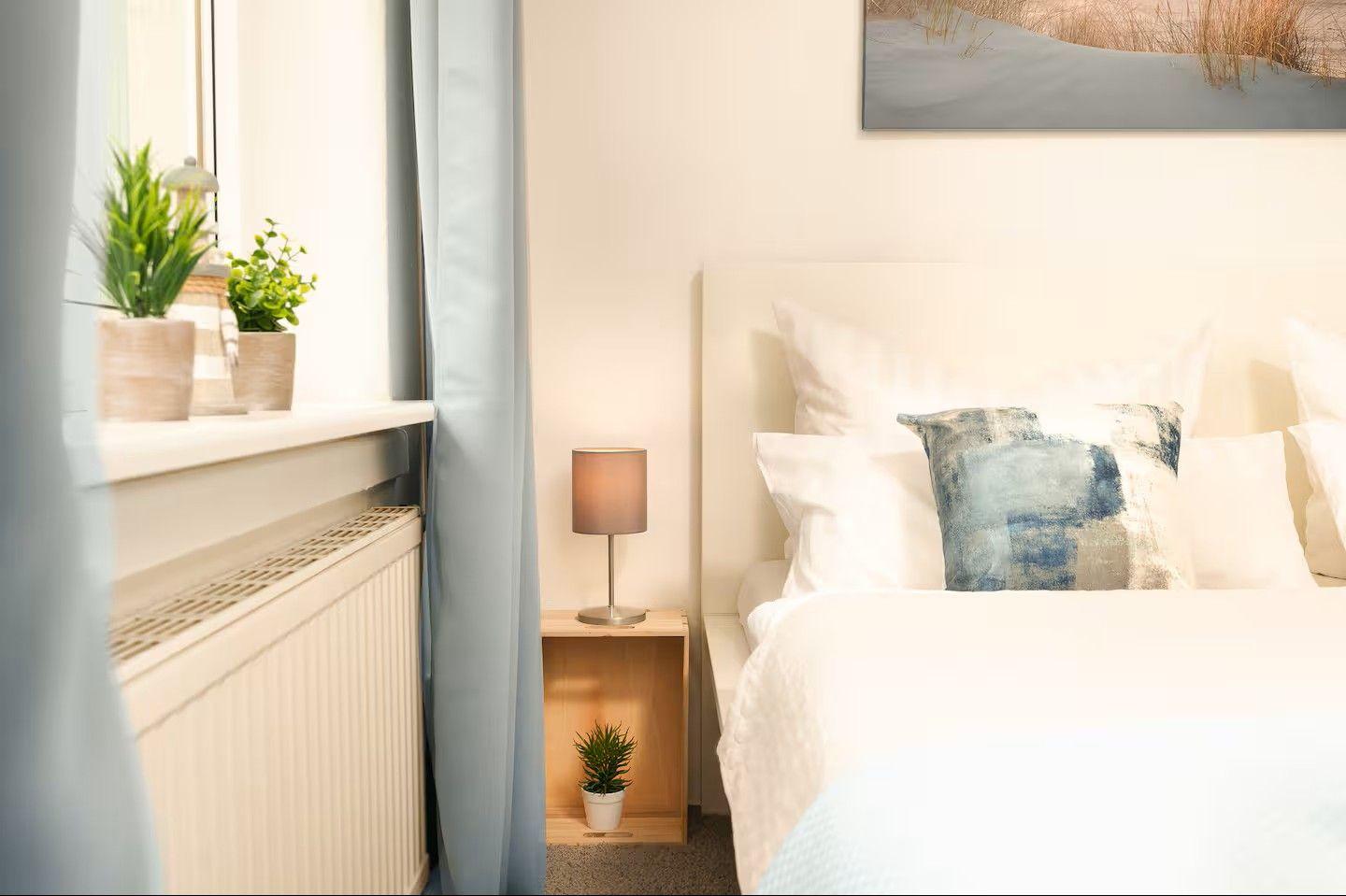 Schlafzimmer mit Bett, Nachttisch, Lampe und Pflanzen an Fensterbank.