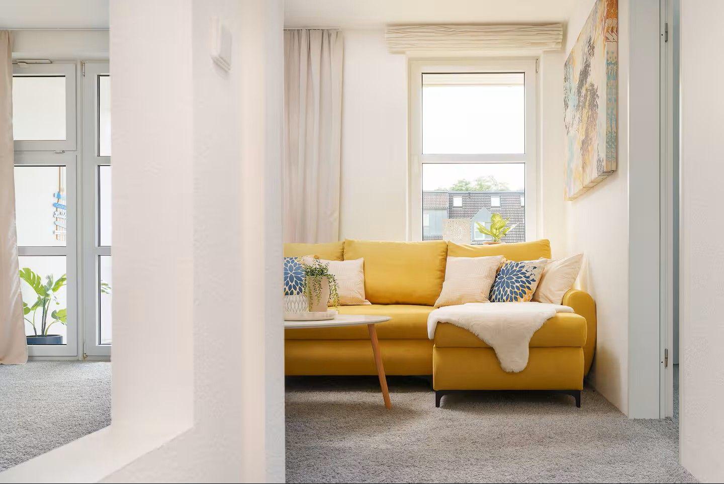Ein gelbes Sofa mit Kissen und Decke steht vor einem Fenster. Links ist eine Glastür zu sehen.
