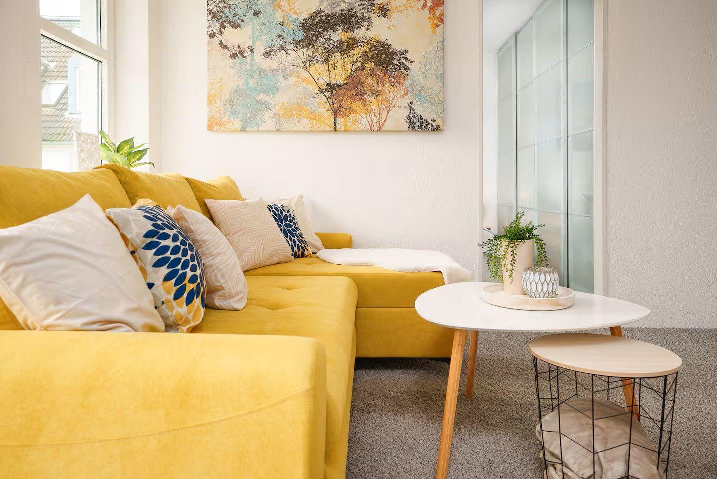 Gelber Sofa mit Kissen, runder Tisch und Wandbild mit Bäumen.