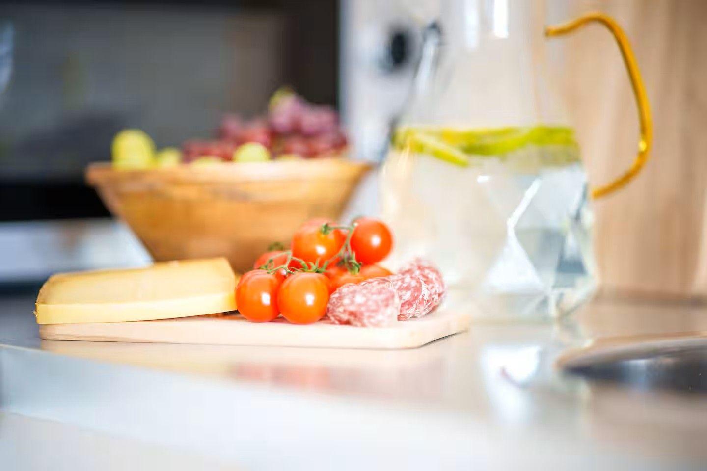 Auf der Arbeitsplatte stehen Käse, Tomaten und Wurst neben einem Wasserglas.