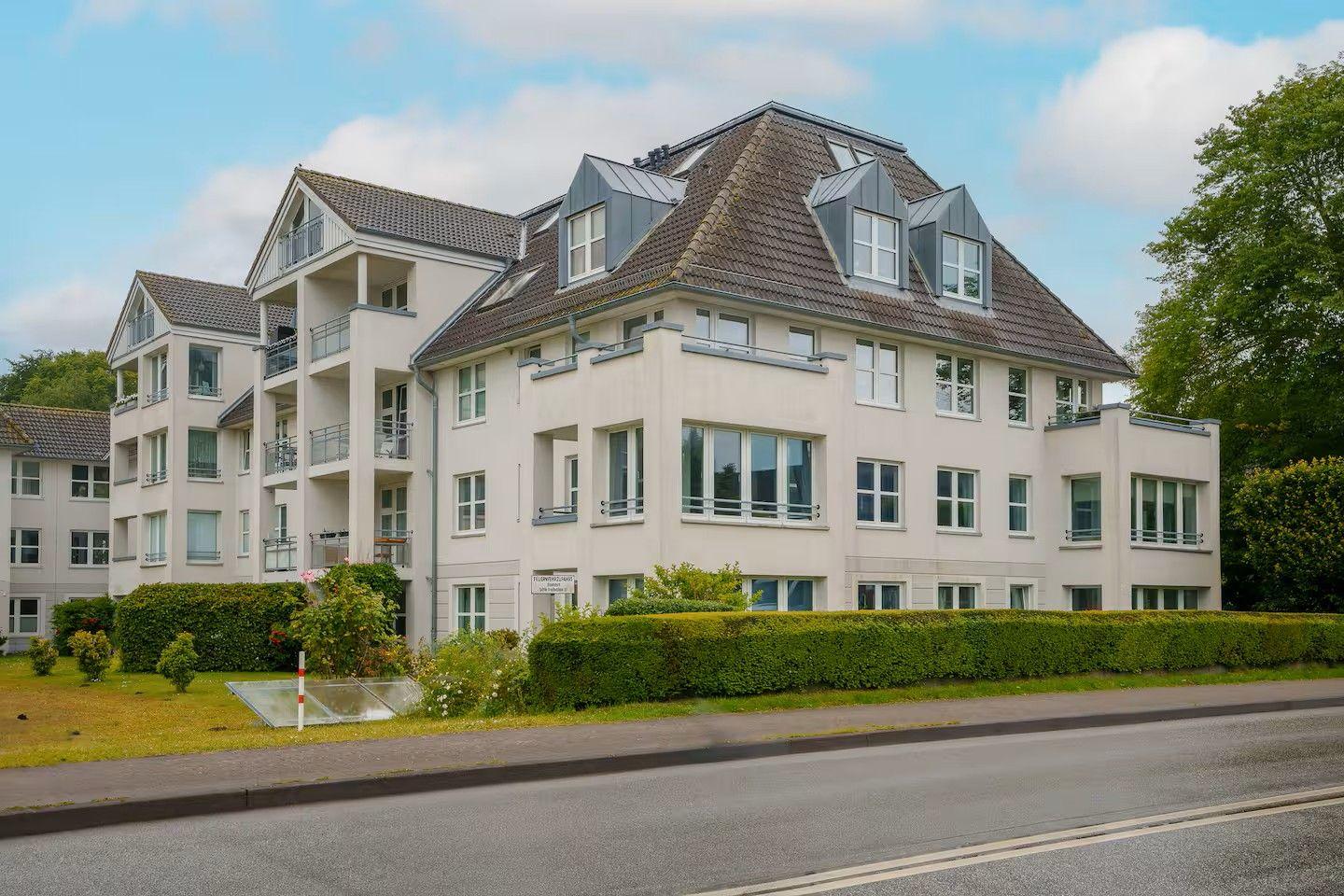 Modernes Mehrfamilienhaus mit Balkonen und Dachgauben. Grünanlagen und Straße im Vordergrund.