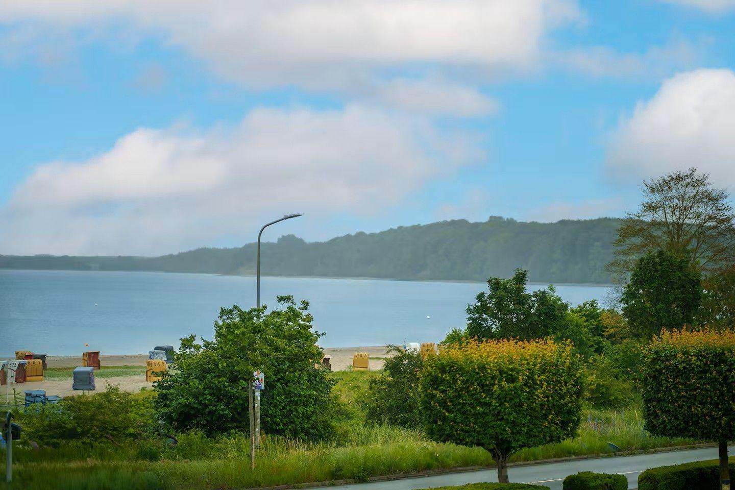Blick auf einen See mit Strand, Bäumen und einem Hügel im Hintergrund.