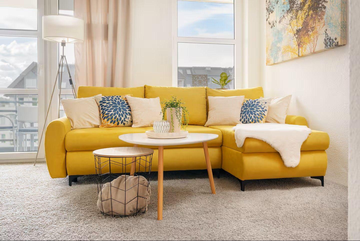 Ein gelber Sofa mit Kissen und Decke vor großen Fenstern.