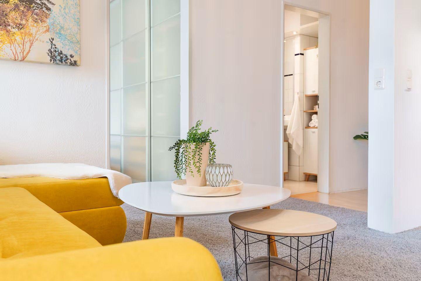 Ein Wohnzimmer mit gelbem Sofa, runden Tischen und einer Pflanze auf dem Tisch.