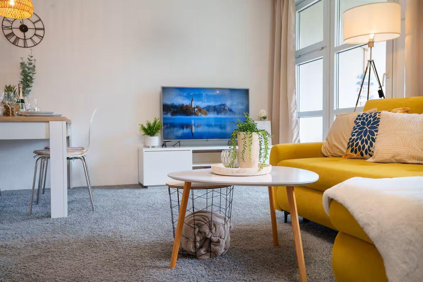 Ein Wohnzimmer mit gelbem Sofa, Fernseher und Esstisch. Pflanzen und Lampen sind vorhanden.