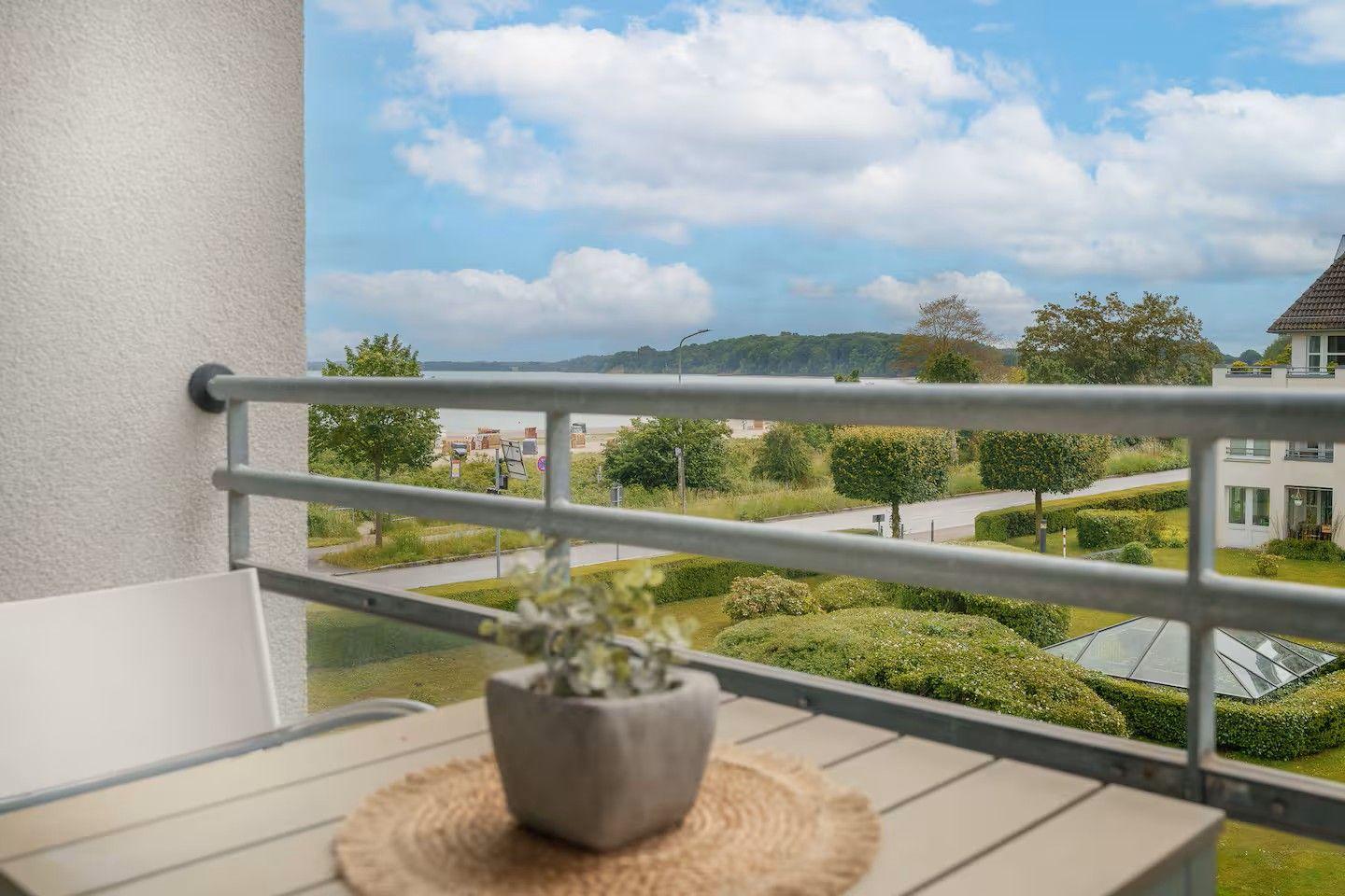 Balkon mit Tisch und Pflanze. Blick auf Grünflächen, Straße und Wasser.