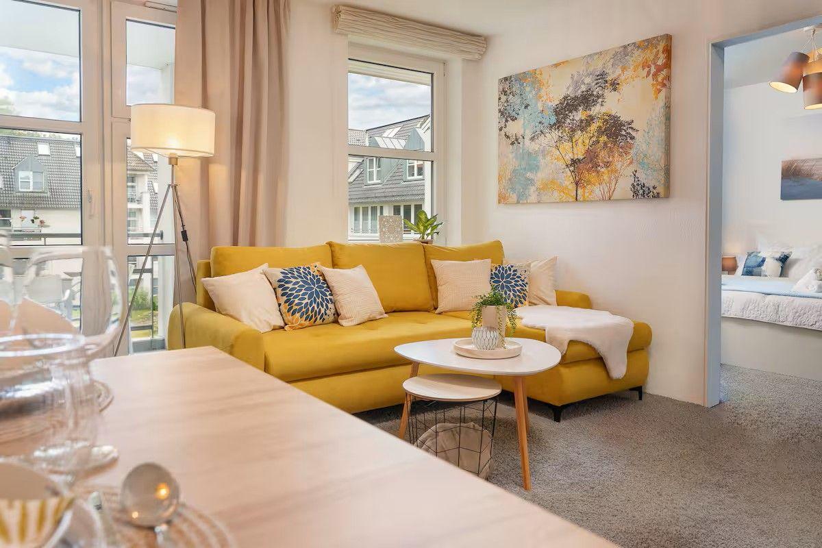 Wohnzimmer mit gelbem Sofa, Tisch und Fenstern. Schlafzimmer im Hintergrund.