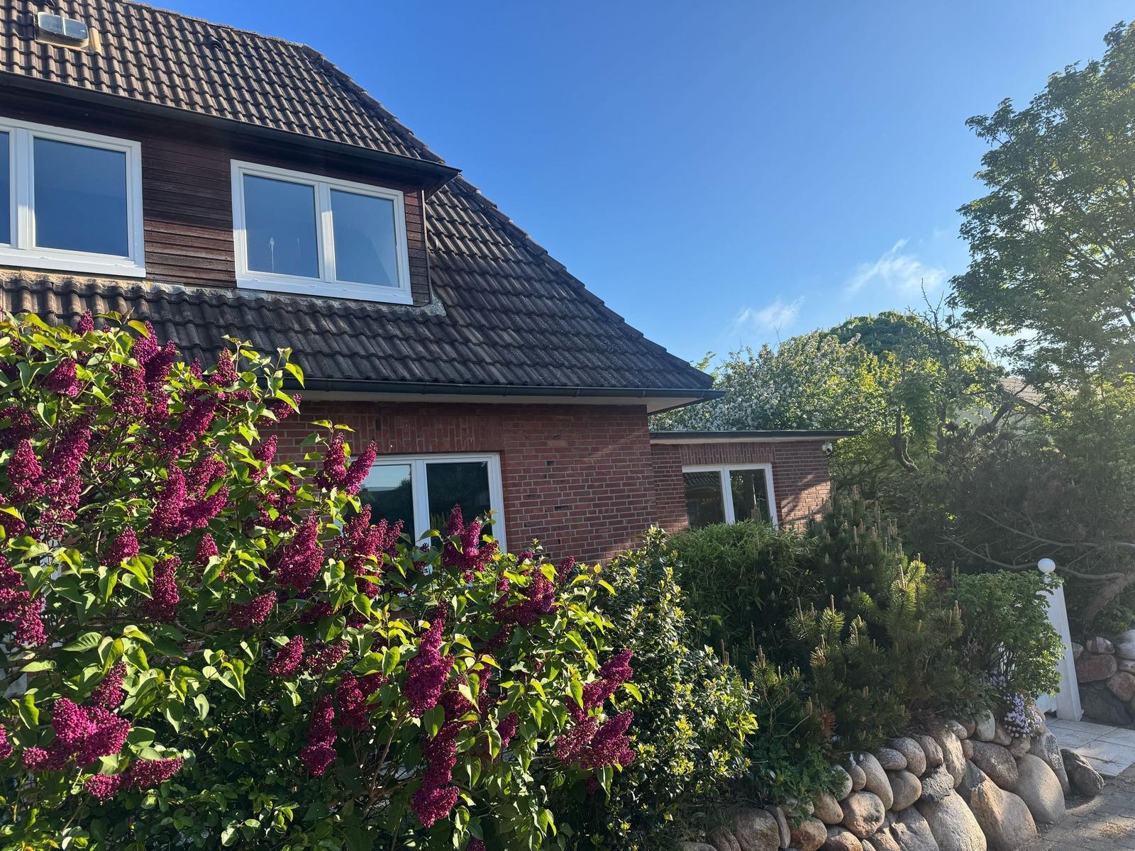 Haus mit Ziegelfassade und Dachgauben, umgeben von Blumen und Bäumen unter blauem Himmel.