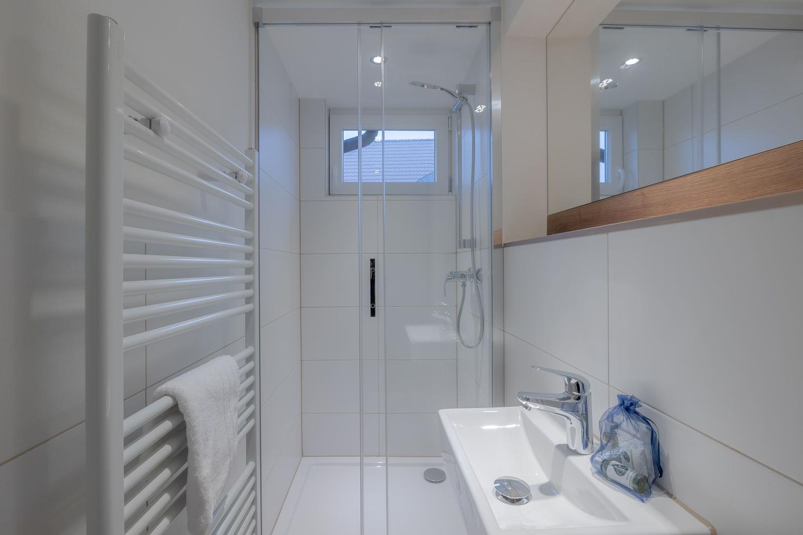 Badezimmer mit Dusche, Waschbecken und Heizkörper. Fenster sorgt für Tageslicht.