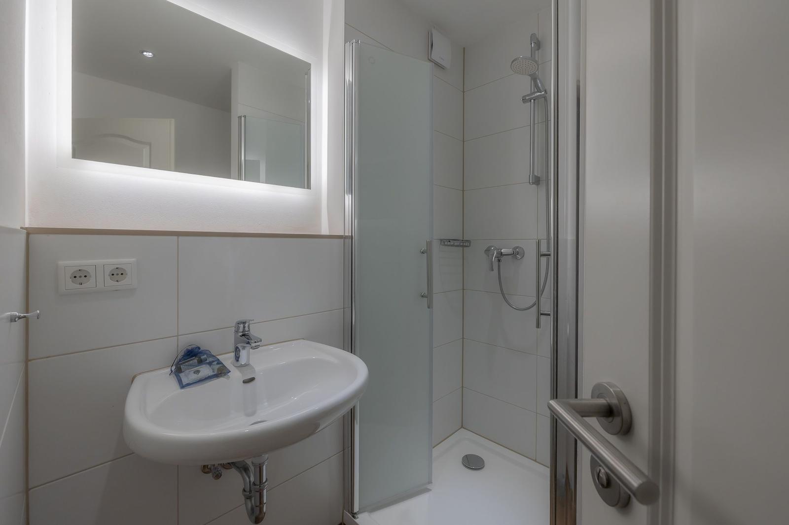 Badezimmer mit Dusche, Waschbecken und beleuchtetem Spiegel.