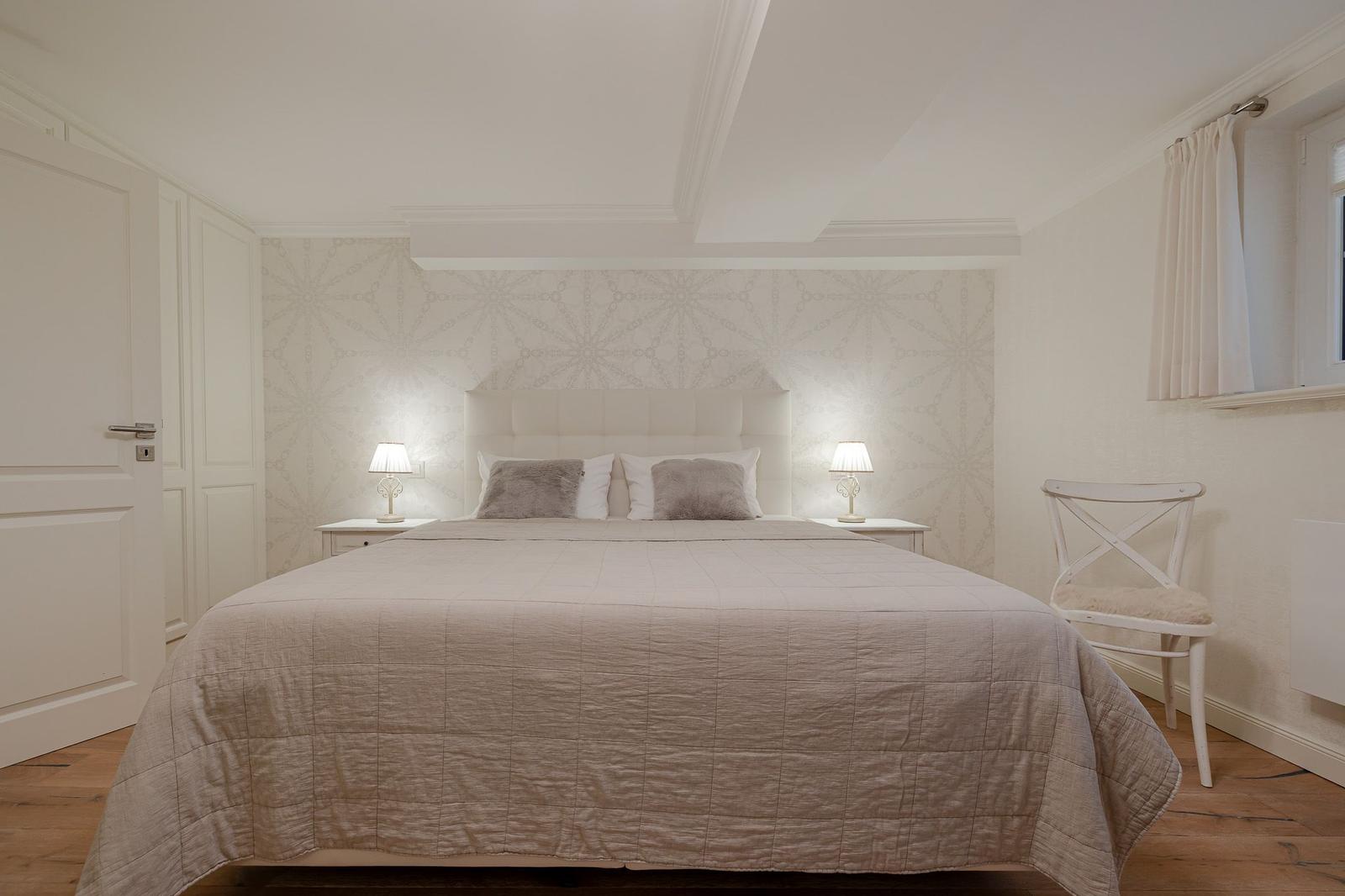 Schlafzimmer mit Bett, Nachttischen und Stuhl. Tapete und Möbel sind weiß.