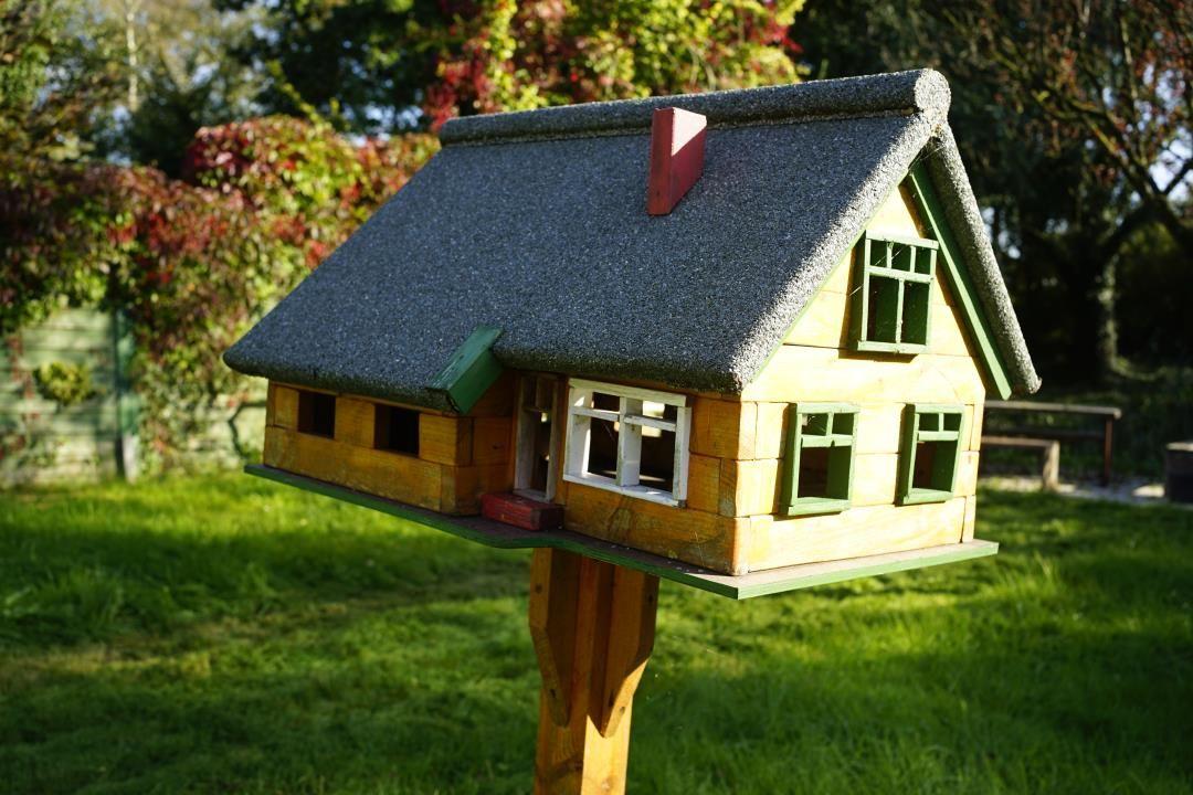 A small wooden house with a gray roof and green window frames stands on a post in the garden.